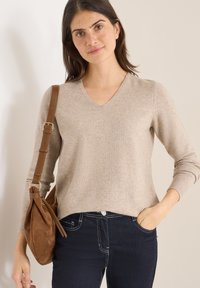 Beżowy sweter w serek z prążkowanym wykończeniem, w połączeniu z ciemnoniebieskimi dżinsami. Brązowa zamszowa torebka na ramię z przekręconym uchwytem i zamkiem błyskawicznym.