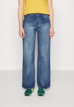 Persona vestida con vaqueros azules de pierna ancha, camiseta amarilla y zapatillas verdes, de pie frente a un fondo blanco liso.