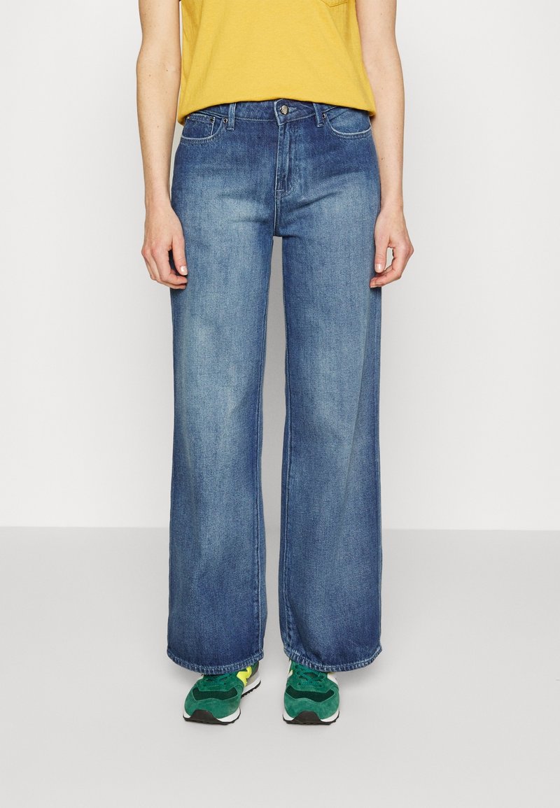Personne portant un jean bleu à jambes larges, un haut jaune et des baskets vertes, debout devant un fond blanc uni.