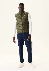 Olijfgroene gewatteerde vest met een hoge kraag en ritssluiting. Gedragen over een witte lange mouwtop, gecombineerd met donkerblauwe broek en witte sneakers.