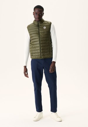 Olive green padded vest with a high collar and zip closure. Worn over a white long-sleeve top, paired with dark navy pants and white sneakers.