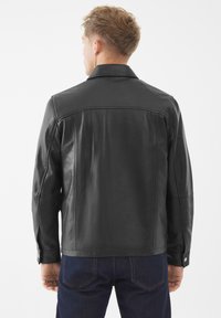 Hombre con una chaqueta de cuero negra y vaqueros azul oscuro, visto desde atrás sobre un fondo blanco liso.