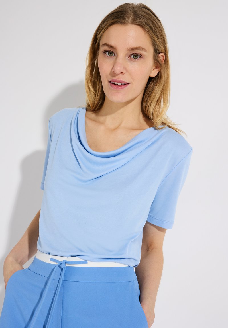 Vrouw met schouderlang blond haar, draagt een lichtblauw korte mouw top met drapering aan de hals en bijpassende blauwe broek met een witte riem.