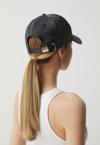 Czarna czapka z błyszczącym wykończeniem, z regulowanym zapięciem z tyłu z metalowym akcentem w kolorze srebrnym. Dopasowany kształt, gładka tekstura.