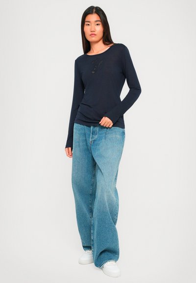 Žena u crnoj dugoj majici s dugim rukavima, širokim svijetloplavim trapericama i bijelim tenisicama, stoji ispred neutralne svijetle pozadine.