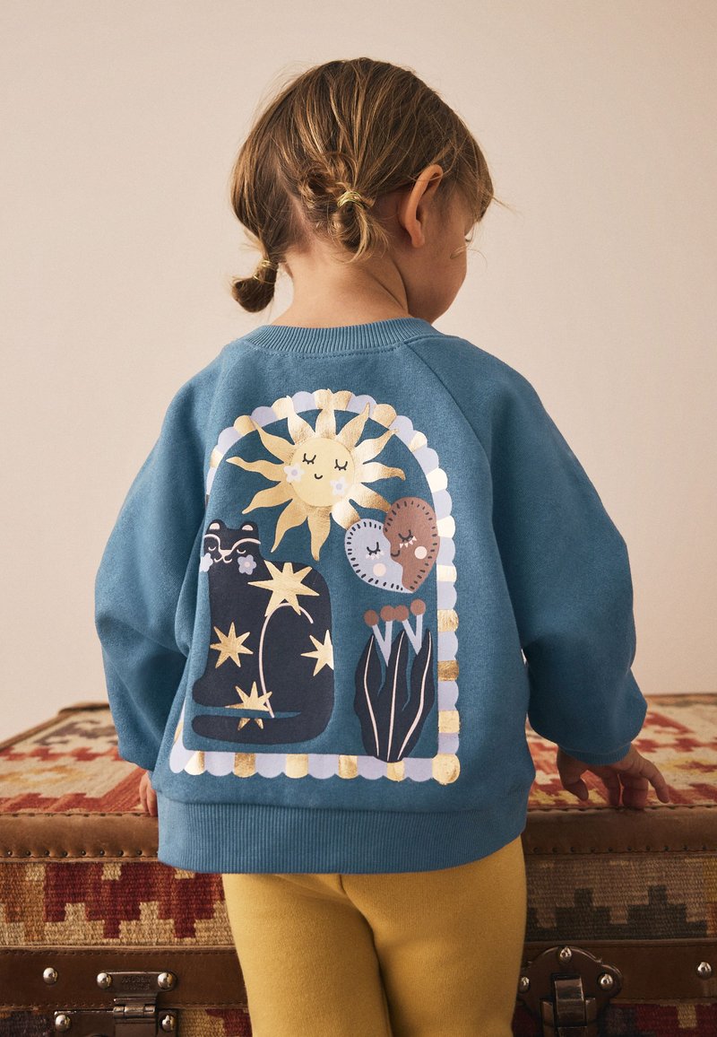 Türkisfarbener Pullover mit großem Motiv einer Sonne, einer Katze, Herzen und Pflanzen auf der Rückseite. Weiche Textur mit gerippten Bündchen an Ärmeln und Saum.