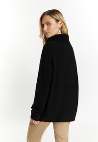 Maglione nero a maglia con colletto alto, polsini a coste e vestibilità ampia, indossato sopra pantaloni beige. La texture appare morbida e grossa.
