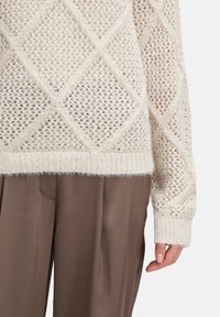 Beige gestrickter Pullover mit Rautenmuster und gerippten Bündchen, kombiniert mit lockeren braunen Hosen, die einen Teil des Oberkörpers und der Hand einer Person zeigen.
