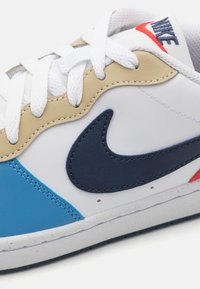 Sneaker with white leather upper, navy swoosh logo, beige accents, and blue overlays. Features white laces and textured rubber sole.