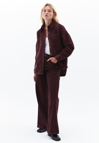 Chemise en velours côtelé bordeaux à boutons avec deux poches poitrine, portée par-dessus un débardeur blanc, associée à un pantalon large en velours côtelé assorti et des chaussures noires.