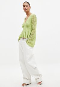 Hellgrübloses Spitzenoberteil mit langen Ärmeln, V-Ausschnitt und Peplum-Taille. Kombiniert mit weiten weißen Hosen und weißen Riemchenschuhen.