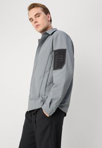 Veste grise à boutons dans un matériau lisse et léger, avec une poche zippée sur le bras gauche, dotée d'un accent noir et d'un logo.