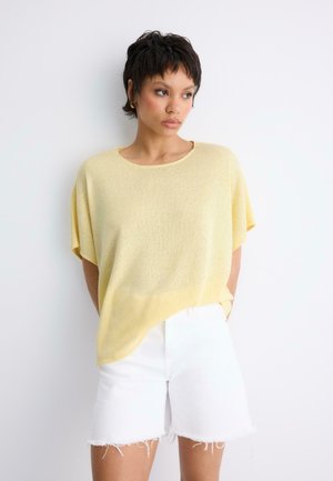 Frau mit kurzen dunklen Haaren, die ein locker sitzendes gelbes Strickoberteil und weiße, ausgefranste Shorts trägt, steht vor einer weißen Wand und schaut nach links.