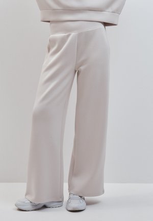 Personne portant un pantalon large beige clair et des baskets blanches, debout devant un fond uni.