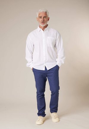 Mann mit weißen Haaren und Bart, der ein weißes Hemd, blaue Hose und beige Turnschuhe trägt, steht mit den Händen in den Taschen.