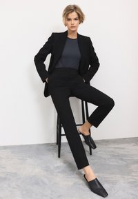 Zwart, op maat gemaakte blazer en broek met een gladde textuur, gecombineerd met een donker topje en zwarte mules, zittend op een zwarte kruk tegen een lichte achtergrond.