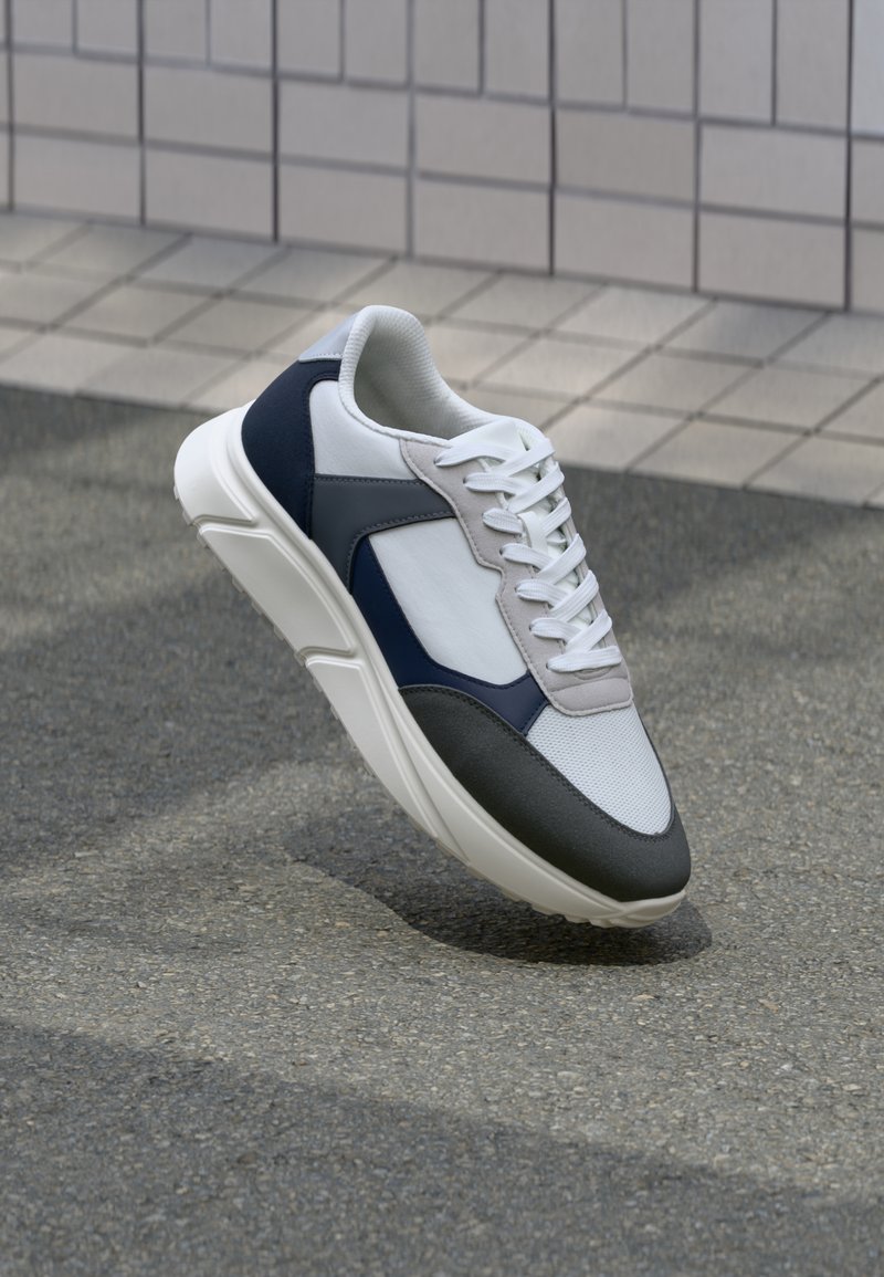 Fehér, tengerészkék és szürke sneaker vegyes anyagokból készült dizájnnal, amely textúrázott felsőrésszel, lapos fűzőkkel és párnázott talppal rendelkezik.