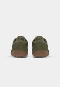 Par tenisica u maslinasto zelenoj boji marke Nike, izrađenih od semiša i mrežastog materijala, prikazanih sa stražnje strane, gdje se vide smeđi gumeni potplati i crni Nike logotip.