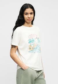 Νεαρή γυναίκα με μακριά σκούρα μαλλιά, που φοράει λευκό T-shirt με πολύχρωμο σχέδιο παραλίας και φοίνικες και ανοιχτό πράσινο παντελόνι.