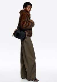 Abrigo de piel sintética marrón con textura a rayas, combinado con pantalones anchos de pierna en oliva y un bolso de nailon negro con borlas de colores. Sandalias marrones.