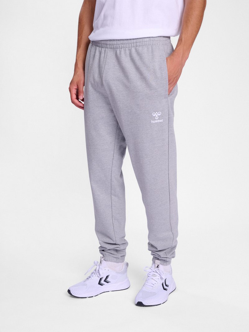 Mann trägt hellgraue Hummel-Sweatpants mit elastischem Bund und Bündchen, kombiniert mit weißen Sportschuhen mit schwarzem Chevron-Logo.