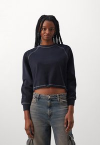 Sudadera recortada de navy en tejido suave con mangas raglán y costuras en contraste. Combinada con pantalones cargo de mezclilla azul claro con bolsillos laterales.