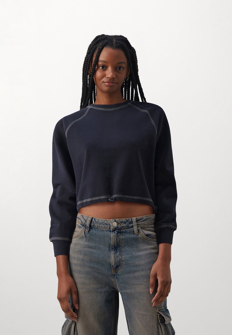 Sudadera recortada de navy en tejido suave con mangas raglán y costuras en contraste. Combinada con pantalones cargo de mezclilla azul claro con bolsillos laterales.