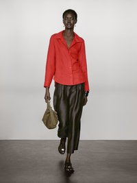Rød linned skjorte med knapper og krave, parret med mørke, blanke wide-leg bukser. Accessories inkluderer en beige taske og sorte sandaler.