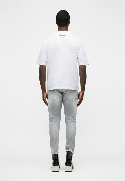Άνδρας που φοράει λευκό πουκάμισο με κοντά μανίκια και λογότυπο "ICONique" στην πλάτη, ανοιχτογκρι τζιν και μαύρες μπότες με τακούνι, στραμμένος με την πλάτη.