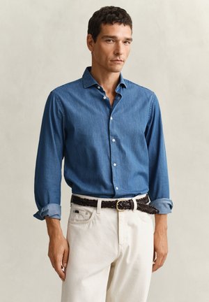 Mann iført blå knappeskjorte med oppbrettede ermer, hvite bukser og et flettet mørkebrunt belte, stående mot en nøytral bakgrunn.