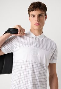 Baltas polo marškinėliai su juodu tinkleliu, dviejų mygtukų apykaklė ir išsiuvinėtas logo. Modelis laiko juodą krepšį, demonstruodamas marškinėlių tinkamumą ir dizainą.