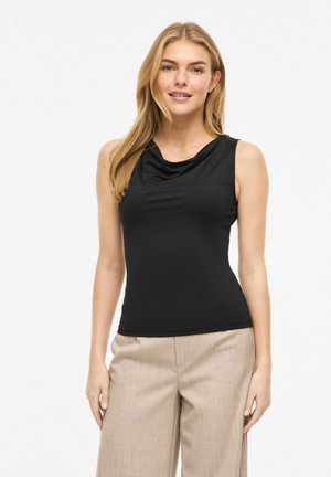 Kvinde iført sort ærmeløs draperet top og beige bukser med vide ben, stående mod en ensfarvet hvid baggrund.