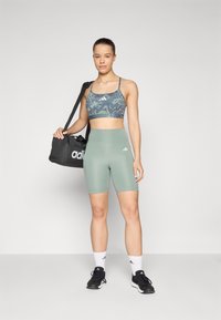 Svart adidas treningsbag, grønn sportsbh med abstrakt mønster, og høyhørte grønne shorts. Modellen har på seg hvite sokker og svarte joggesko.