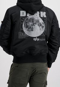 Musta bomber-takki, jossa on joustavat hihansuut ja helma. Takana suuri kuun kuva ja teksti "DARK SIDE OF THE MOON".