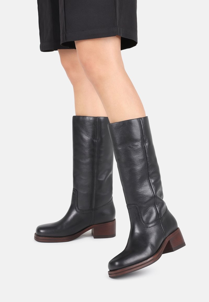 Bottes en cuir noir montantes avec un talon en bois légèrement épais, texture lisse et détails cousus le long des côtés.