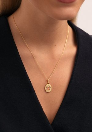 Collarín de oro con un diseño de araña; intrincados detalles en un charm ovalado; cadena fina con una textura suave; llevado sobre una blusa oscura.