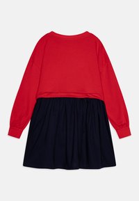 Bluză roșie cu mânecă lungă și guler rotund, asociată cu o fustă bleumarin. Bluza are un design scurt, iar fusta are o textură plisată.