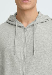 Sweat à capuche gris avec fermeture éclair, tissu texturé, cordons de serrage, doublure dans la capuche, fermeture centrale par zip et détails de couture subtils.