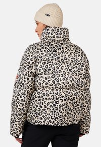 Kurtka puchowa w beżowym kolorze z czarnym wzorem w panterkę. Posiada wysoki kołnierz i pikowaną teksturę, zestawiona z dzianą beżową czapką.