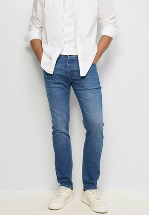 Hombre con zapatillas blancas, vaqueros azules, camisa blanca con las mangas remangadas, de pie con las manos en los bolsillos contra una pared blanca.
