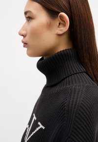 Maglione a collo alto grigio scuro a costine, con un design del logo bianco. Vista ravvicinata laterale che mette in evidenza la texture e i dettagli del colletto.