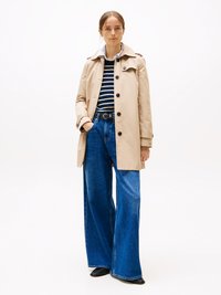 Trench coat beige con chiusura a bottoni, colletto rialzato ed epaulette, abbinato a una camicia a righe, jeans blu a gamba larga e scarpe nere.