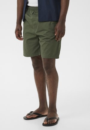 Mand iført olivengrønne shorts, navyblå skjorte og sorte flip-flops, stående mod en ensartet lys baggrund.
