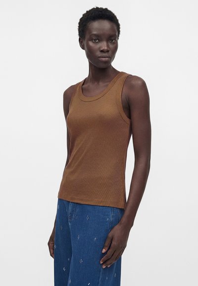 Γυναίκα με σκουρόχρωμο δέρμα και κοντά, σγουρά μαλλιά που φοράει καφέ αμάνικη ραβδωτή μπλούζα και μπλε τζιν με μικρά λευκά σχέδια, στέκεται μπροστά σε λευκό φόντο.