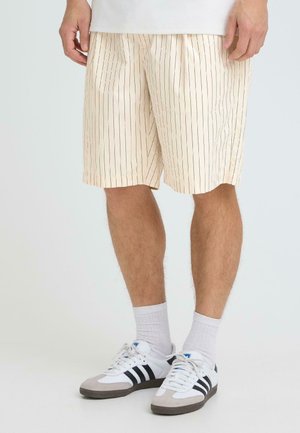 Beine mit beigen Shorts mit dünnen schwarzen Streifen, weißen kurzen Socken und weißen Turnschuhen mit schwarzen Streifen vor einem schlichten Hintergrund.