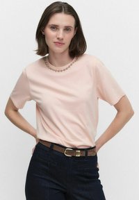 Kortärmad persikofärgad T-shirt med rund halsringning. Kragen har en blommig pärldesign. Bärs tillsammans med mörka jeans och ett brunt bälte.