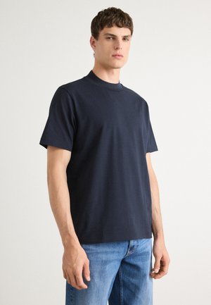 Marineblaue Baumwoll-T-Shirt mit hohem Kragen und kurzen Ärmeln, das ein Markenlogo am Ausschnitt trägt. Getragen mit hellblauen Jeans.