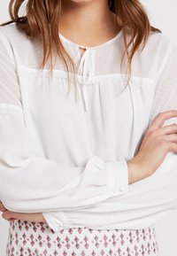 Blusa bianca con maniche lunghe, scollo a cravatta, tessuto trasparente a pois nella parte superiore e dettagli in pizzo. Polsini con bottoni e dettaglio arricciato.
