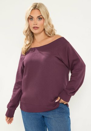 Blond kvinde iført en lilla skulderfri sweater og blå jeans står mod en ensfarvet lys baggrund.