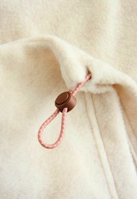 Fleece materiaal in lichtbeige met een gladde textuur. Beschikt over een roze koord met een bruine plastic stopper voor aanpassing.